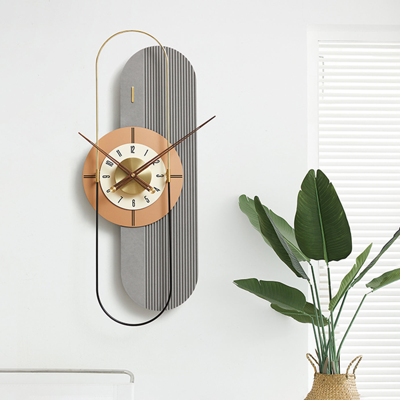 Modern vertical wall clock with a decorative design on a white wall with a plant in the corner.
