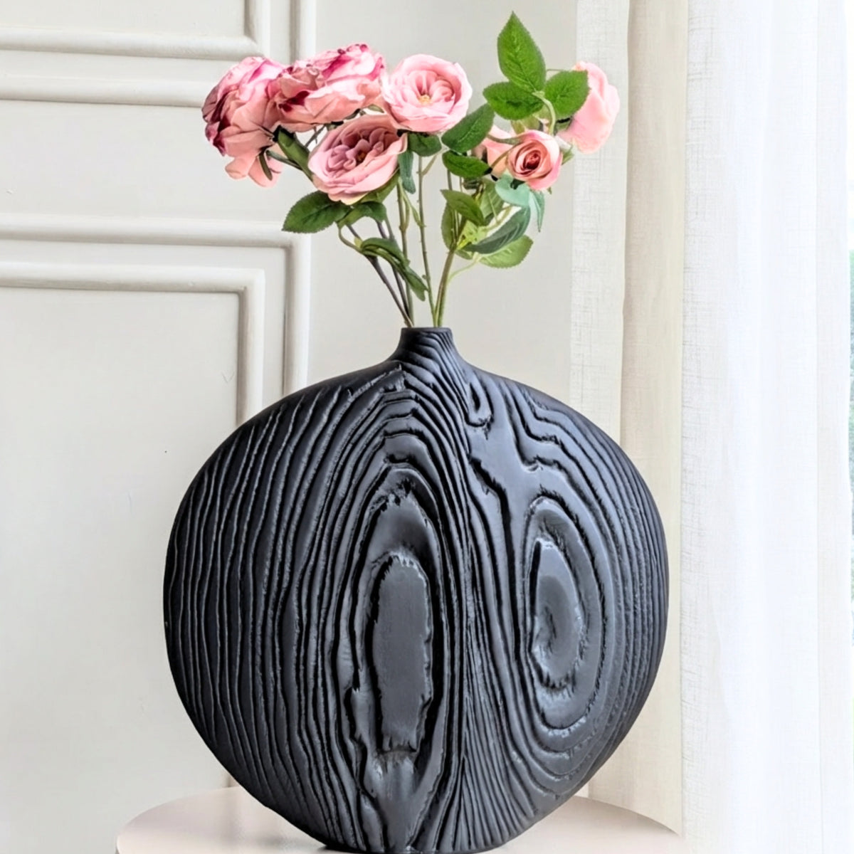 Black textured round ceramic vase styled with pink roses against white wall paneling